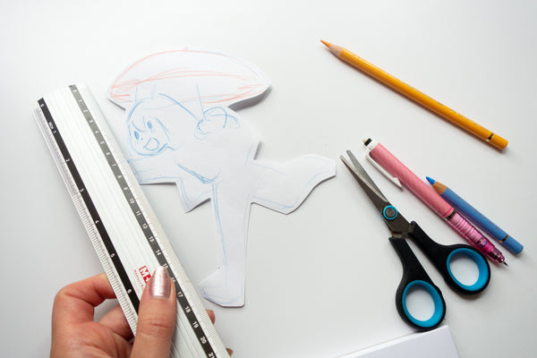Hand holding a ruler over a paper cutout with scissors, pencils, and markers on a white background - Making of image 2