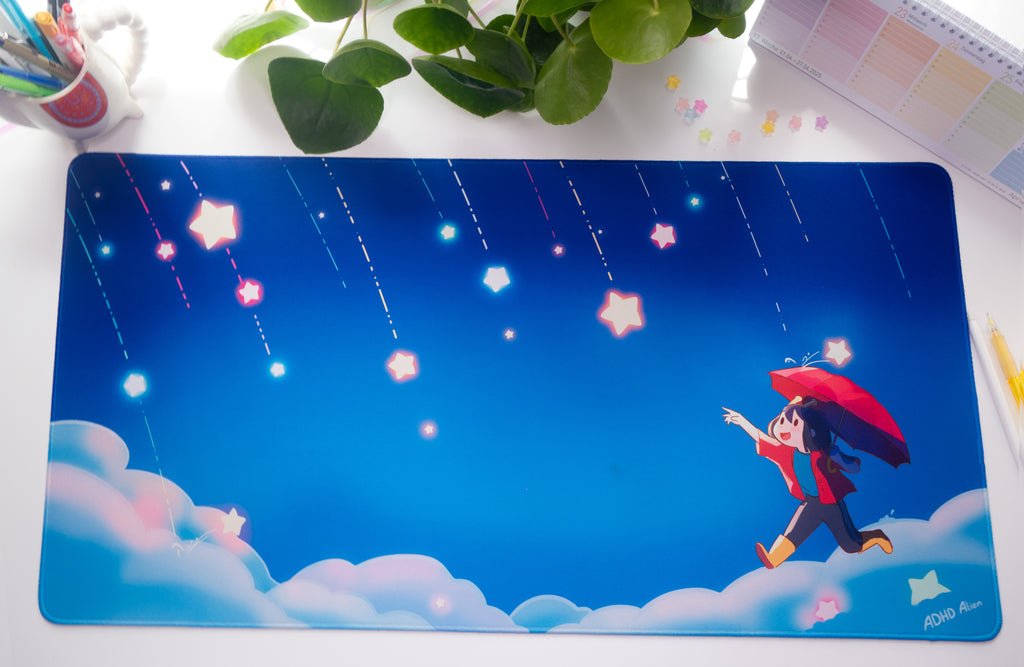 Illustrated desk pad with a character holding an umbrella under a starry sky, placed on a desk with stationery items.