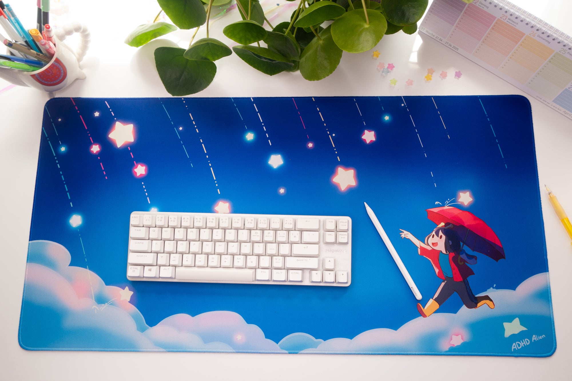 Blue desk mat with ADHD Alien character holding an umbrella. There are a keyboard, and stationery items on the desk.