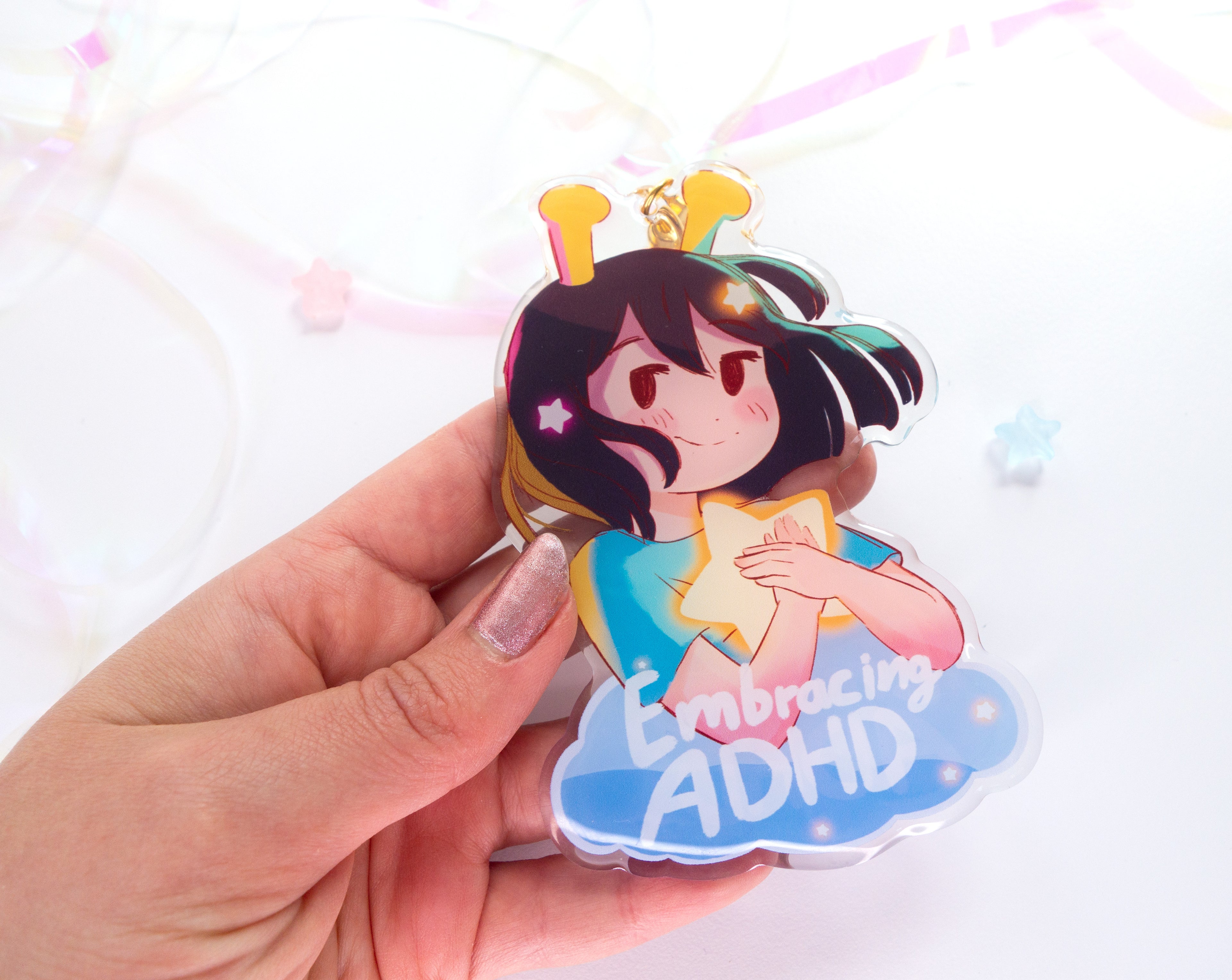 Acrylic charm with an illustration of the ADHD Alien character holding a star, with text 'Embracing ADHD', held by a hand against a light background.