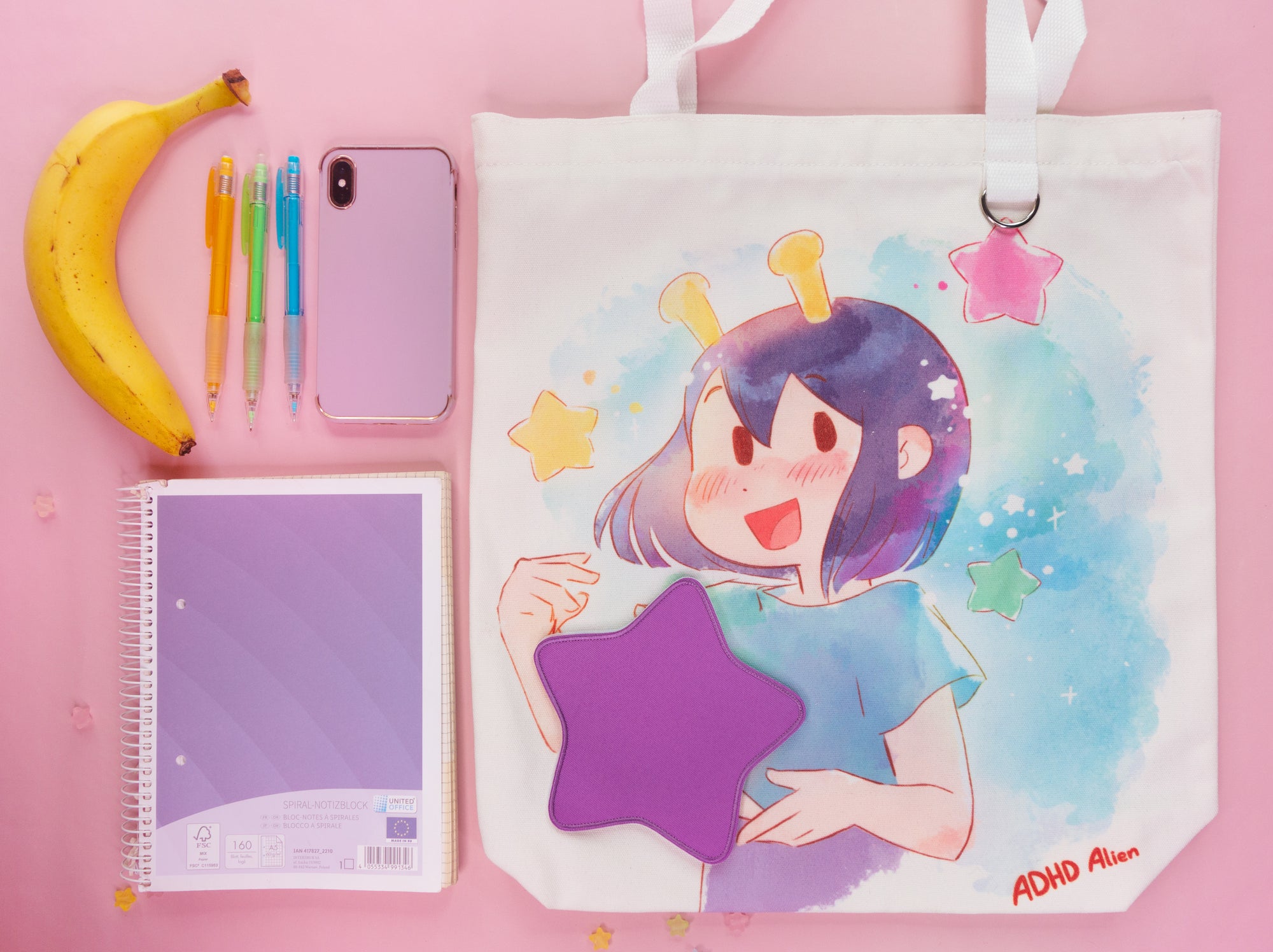 Tote bag with a ADHD Alien holding a star. notebook, phone, and stationery items on a pink background.